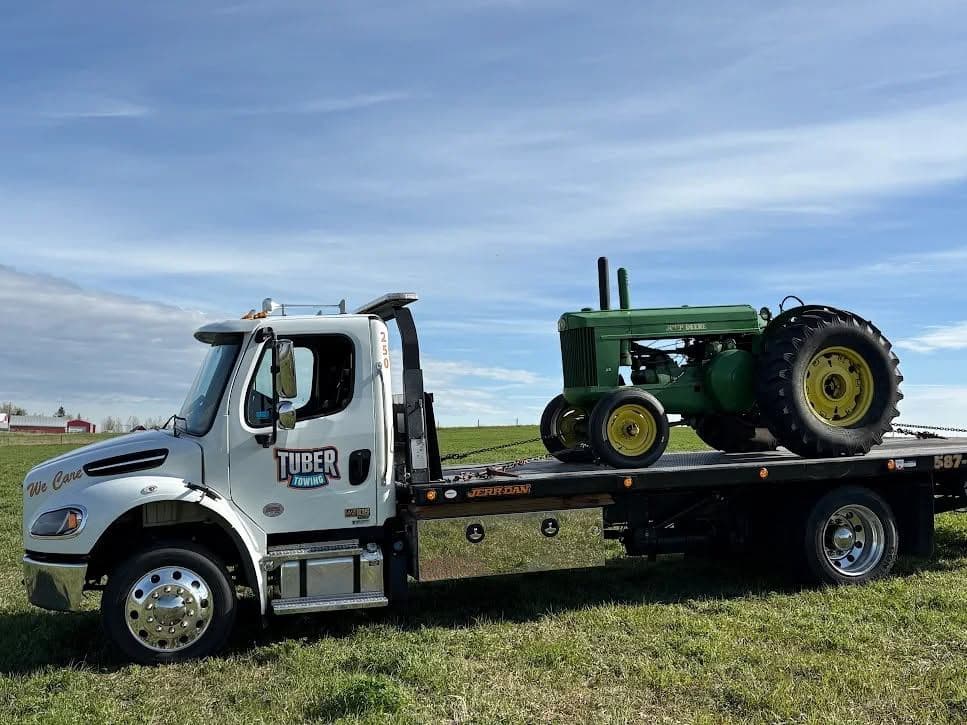 A white Tuber Towing flat deck with "We Care" lettering on the door, carrying a restored green John Deere tractor, parked on grass beside a farm.