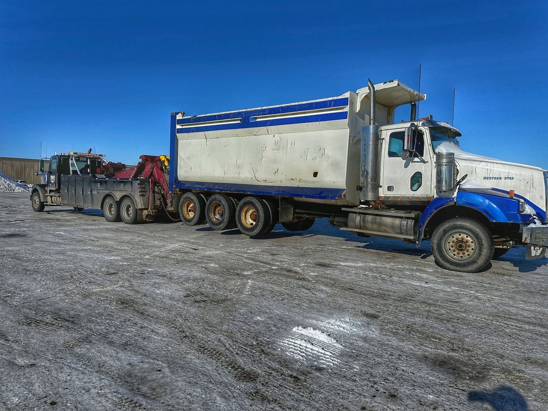 A Tuber Towing heavy wrecker towing a loaded Western Star gravel truck across packed snow under a clear blue winter sky.