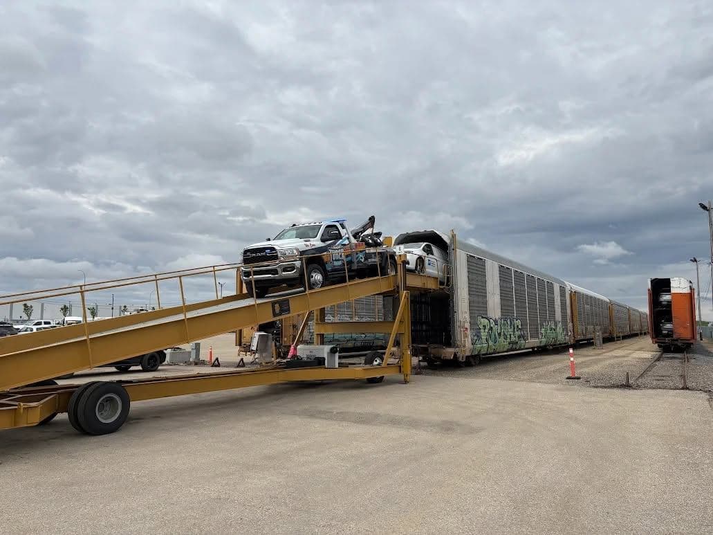 A Tuber Towing service truck driven up a yellow loading ramp onto the upper deck of a rail autorack car, with a line of railcars stretching behind it.